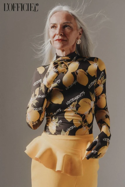 Elegant older woman poses in a black top with lemon print and a yellow skirt in a fashion editorial studio shot. 