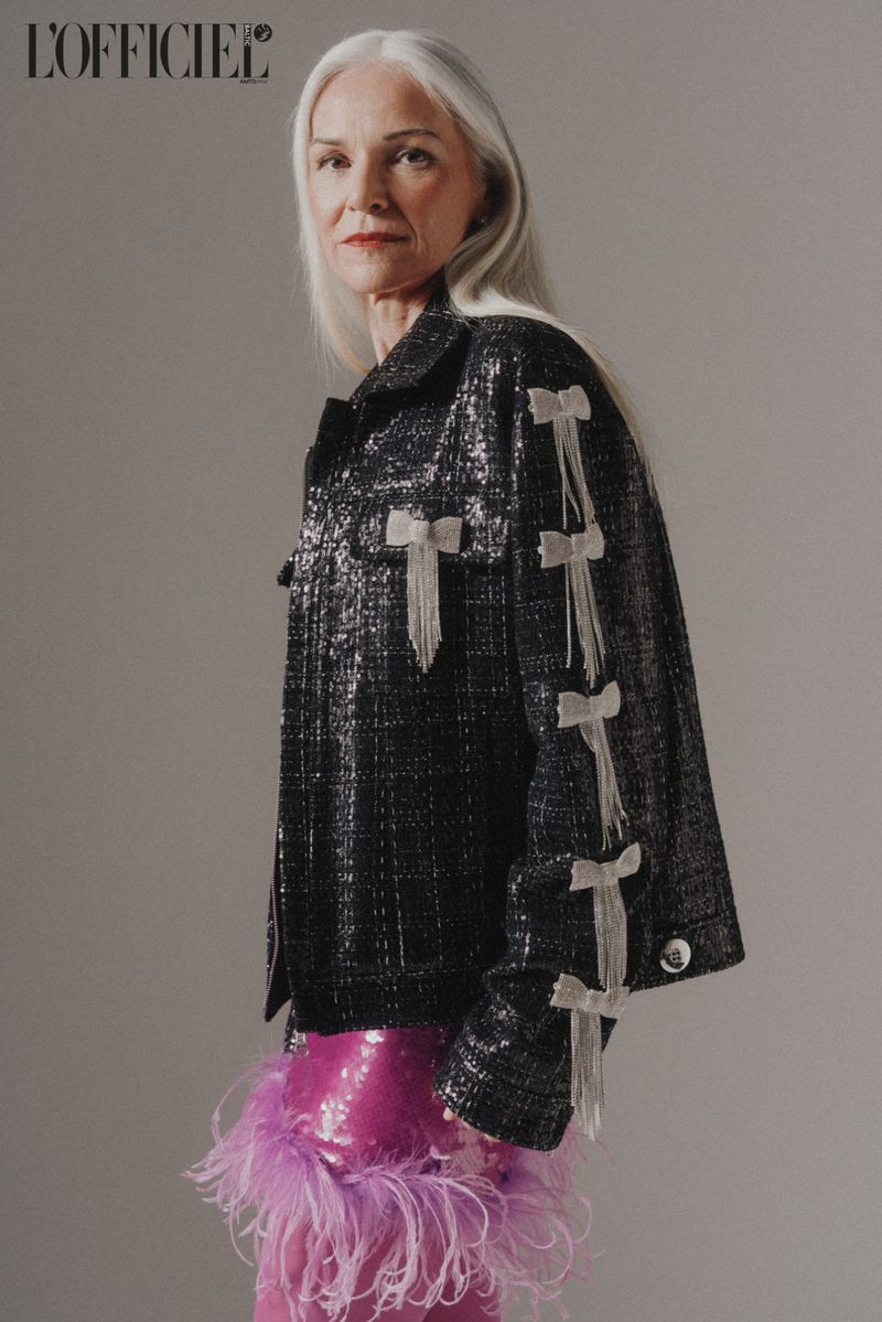 Elegant senior woman in a black tweed jacket with white bow details and a short pink feathered skirt poses in a fashion studio.