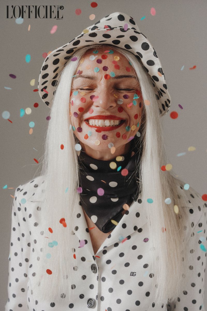 A woman smiles with confetti on her face, wearing a polka dot hat and jacket, with a black and white patterned scarf.