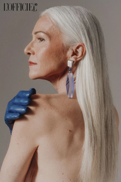 Elegant older woman with long silver hair poses in a beauty shot, showcasing statement earrings and blue leather gloves against a grey background.