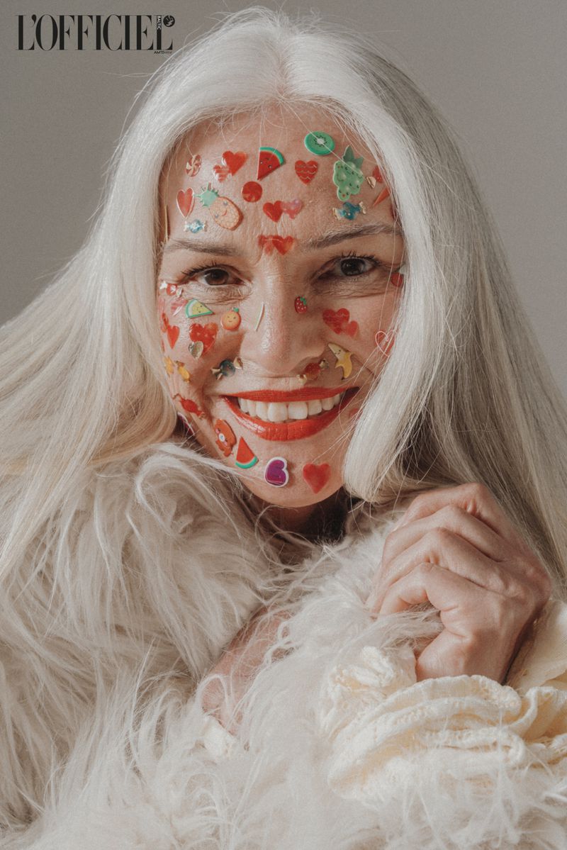 Close-up studio shot of a smiling elderly woman with white hair, wearing a fur-like coat and fruit/heart stickers on her face.