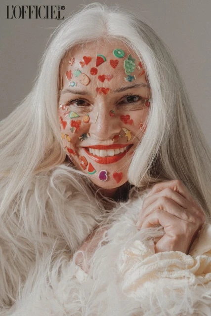 Close-up studio shot of a smiling elderly woman with white hair, wearing a fur-like coat and fruit/heart stickers on her face.
