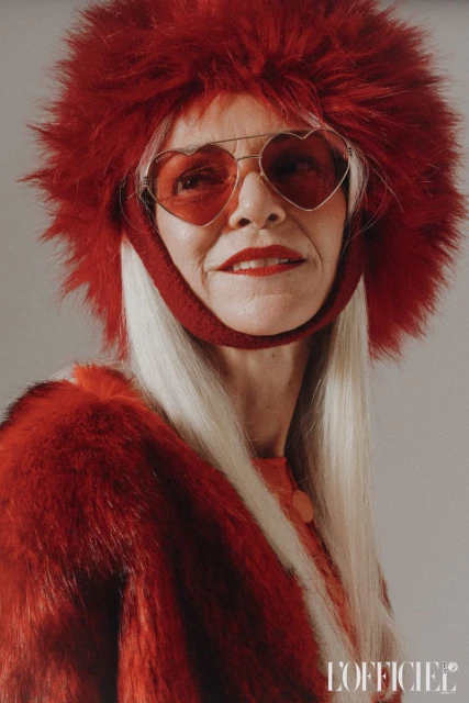 Elderly woman in a red fur coat and hat with heart-shaped sunglasses, posing indoors for a fashion editorial portrait.