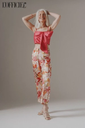 Elderly woman with white hair smiling in a pink crop top and floral trousers against a neutral grey seamless background.