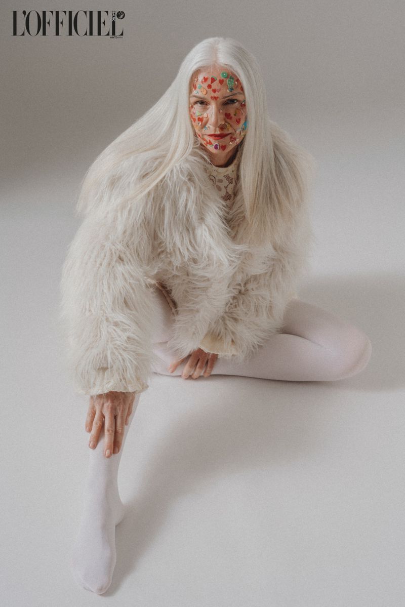 A woman with white hair sits in a studio with colorful heart designs on her face, wearing white tights and a shaggy white coat.