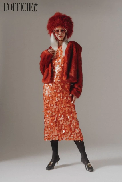 Full-length portrait of a model in a red faux fur hat and jacket, and a sequin dress, posing in front of a neutral backdrop.