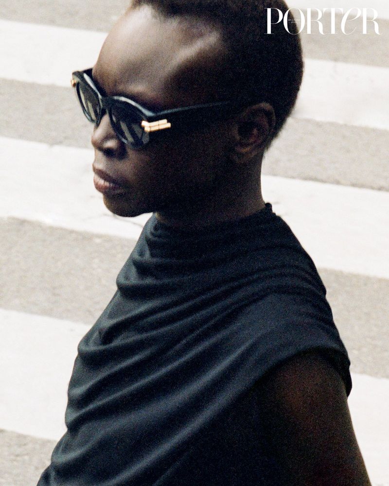 An adult woman wearing sunglasses and a black top poses in an urban setting with light from above, fashion editorial image.