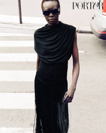 Fashion editorial shot of a Black woman in a draped sleeveless black dress and sunglasses, walking on a crosswalk with a car visible.