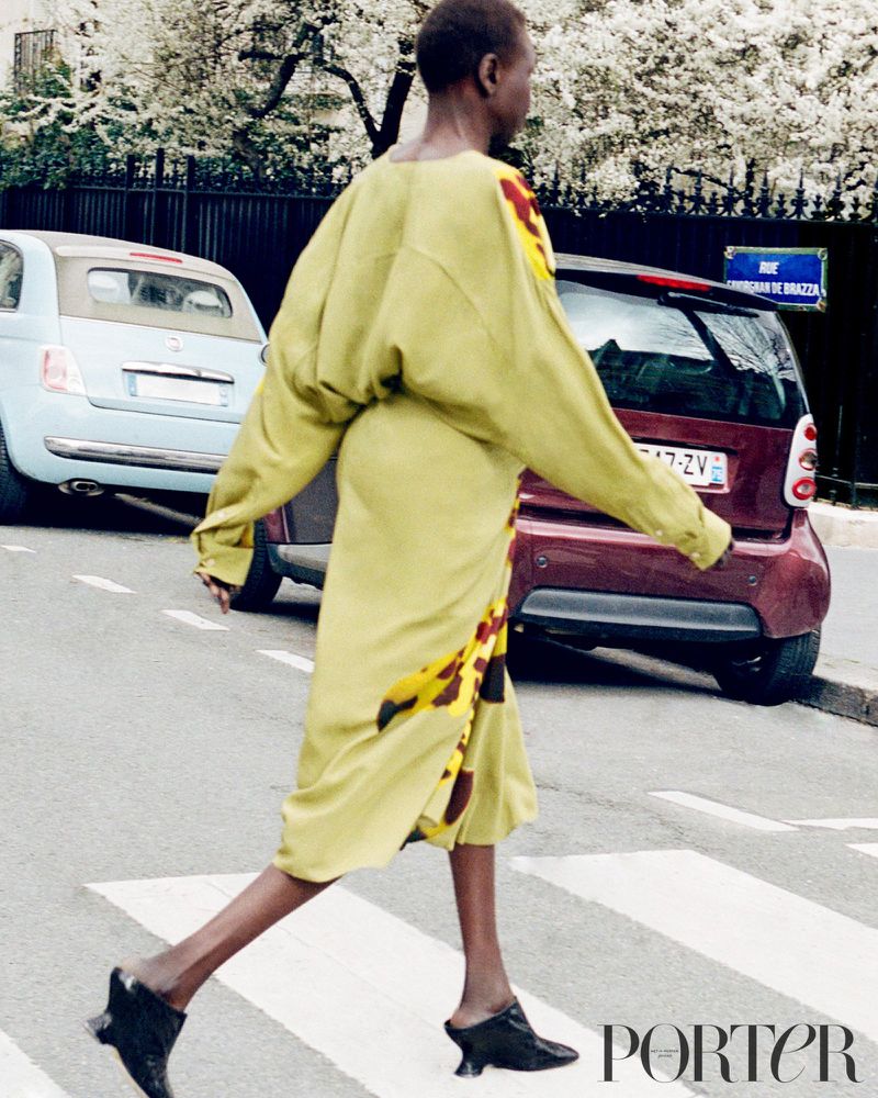 Fashion editorial shot of a Black woman in a light green dress crossing a street with cars and buildings in the background on a sunny day.