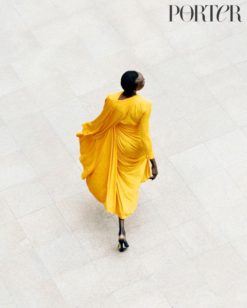 Overhead shot of a Black female in a yellow dress walking on a tile floor, fashion editorial, magazine cover, neutral backdrop, Porter magazine.