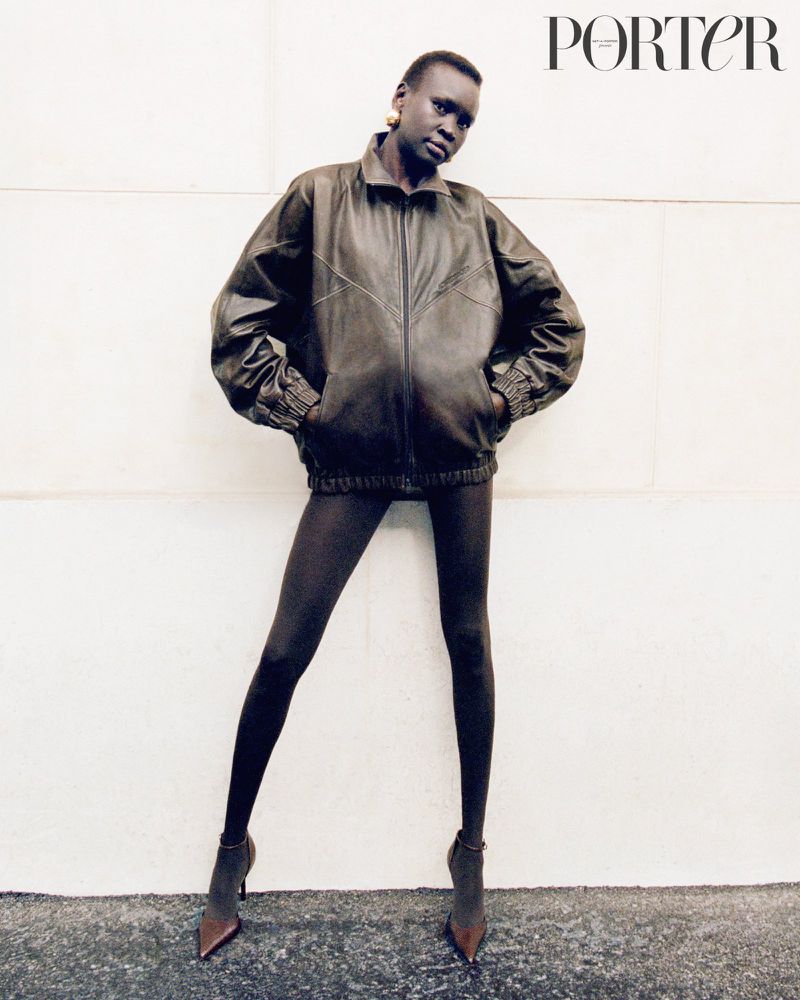 Studio shot of a slender African-descent woman in a leather jacket, tights, and heels against a white wall with the Porter magazine logo.