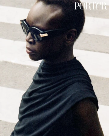 Alek Wek wearing dark sunglasses and a black top against a striped background for Porter Magazine editorial