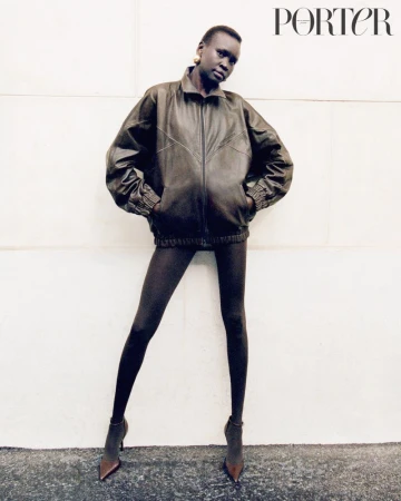 Model Alek Wek in an oversized dark leather jacket and dark tights against a plain wall for Porter Magazine editorial.