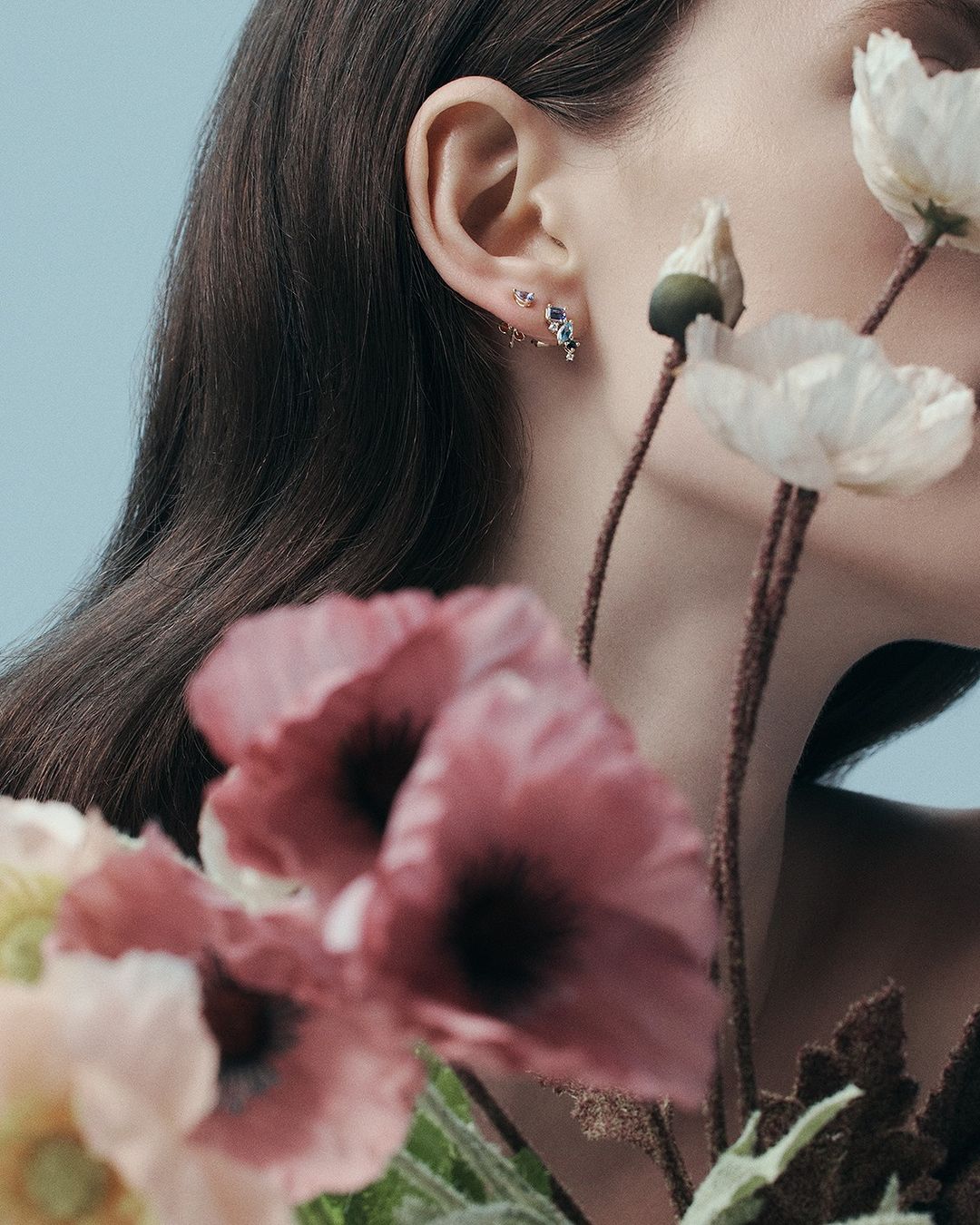 Elegant jewelry advertisement featuring a woman's ear adorned with multiple earrings, accented by a bouquet of pink and white flowers.