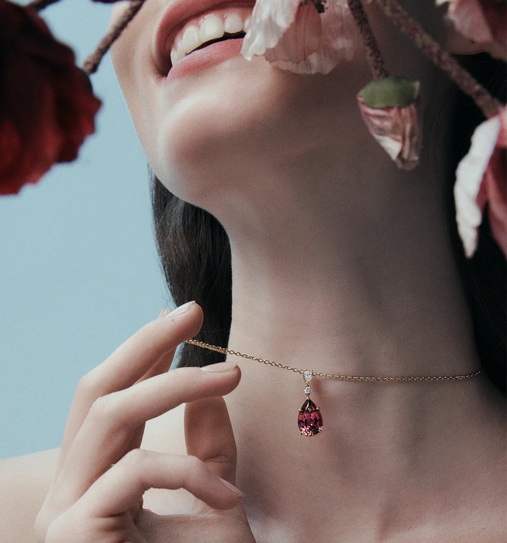 Close-up of a woman wearing a delicate gold chain necklace with a pink tourmaline teardrop pendant against a light blue backdrop.