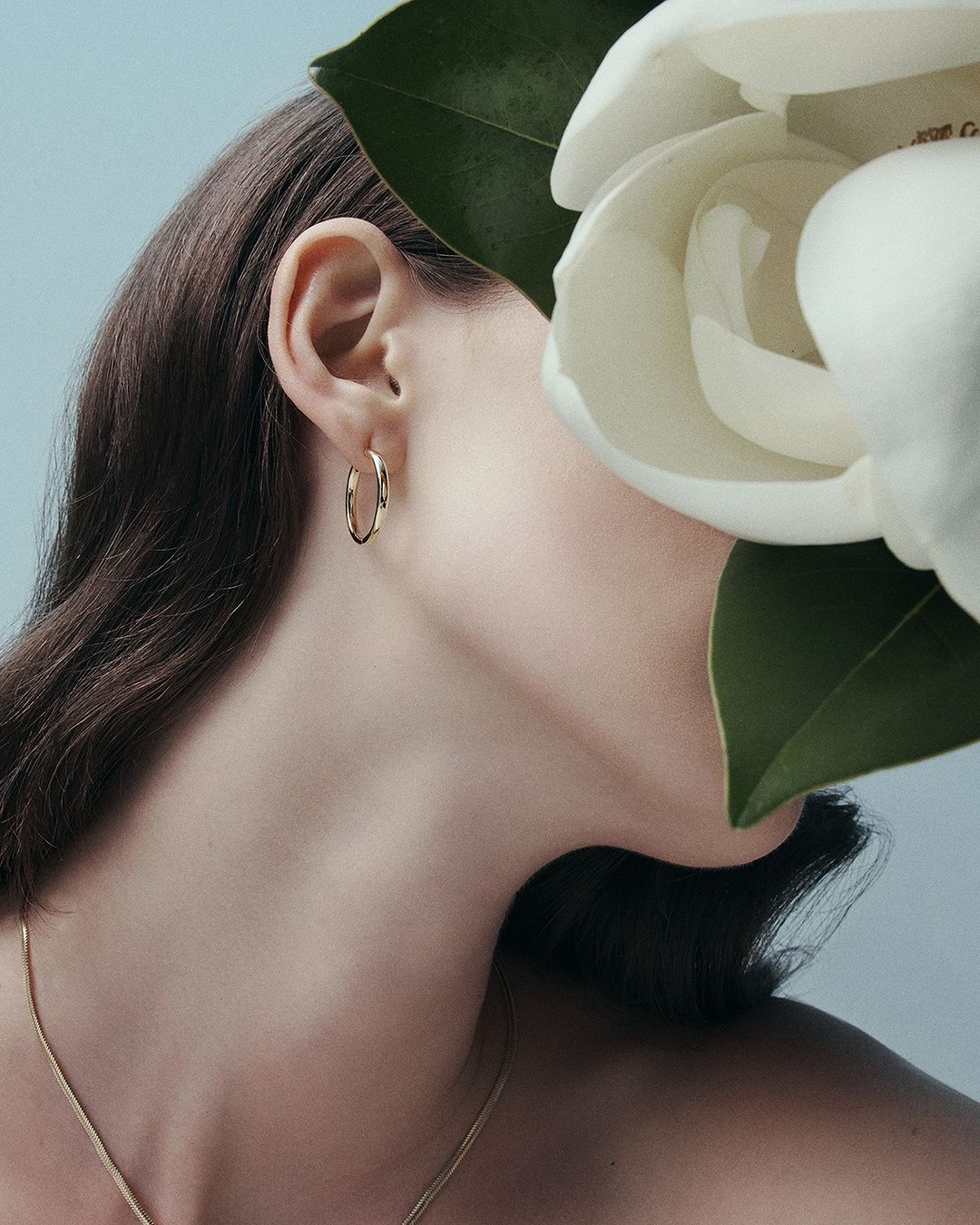 Close-up of a woman with dark hair and gold hoop earring, partially obscured by a white magnolia flower against a light blue background.
