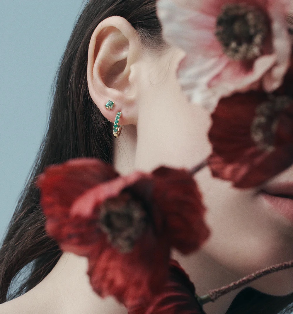 Close-up shot of a person's ear with emerald and gold hoop earrings and a stud earring, flowers visible in the foreground.