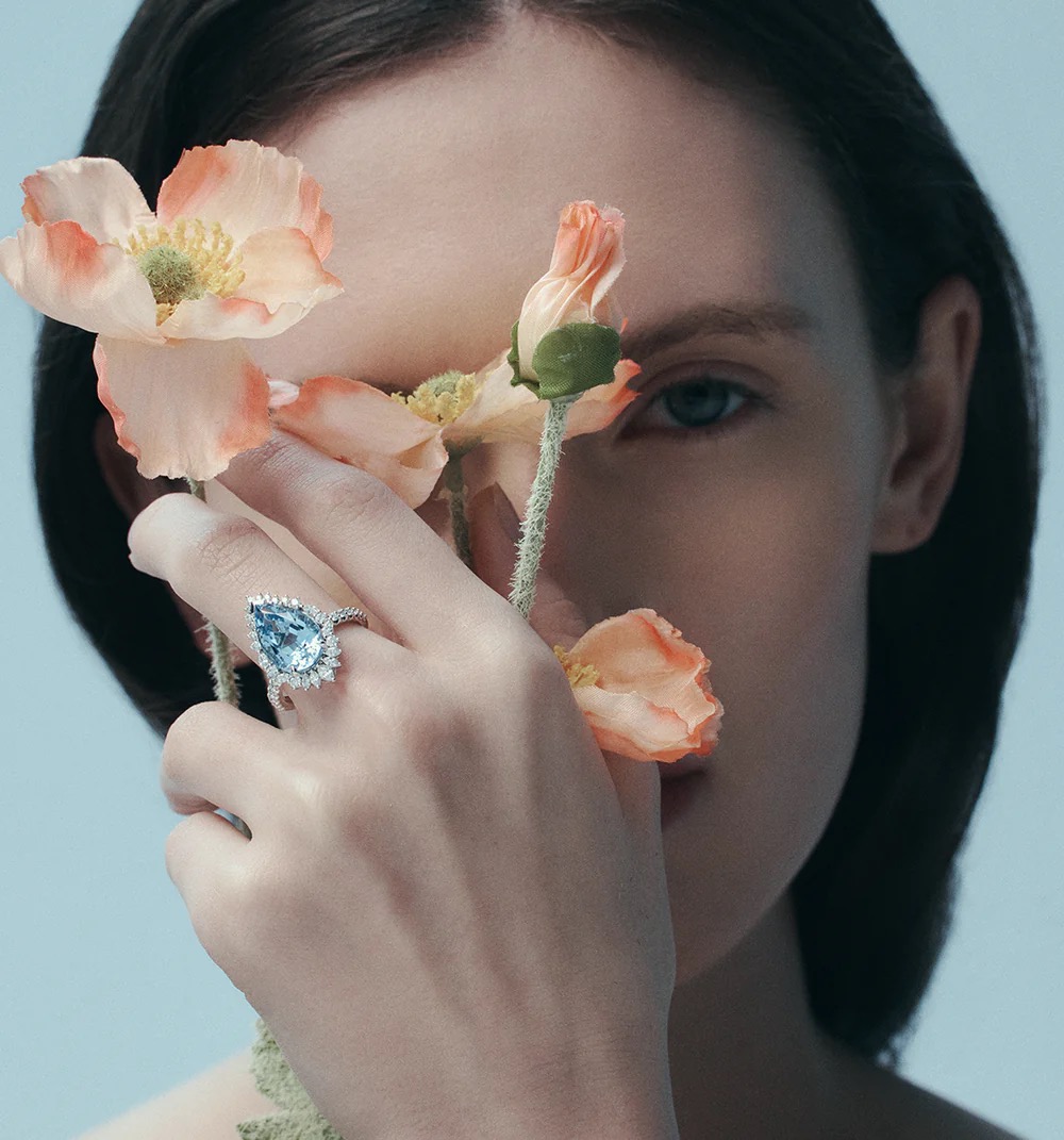 Elegant studio portrait of a woman's hand showcasing a blue gemstone ring, accented by peach flowers against a soft blue backdrop.