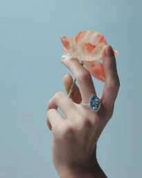 Close-up of a hand wearing a pear-shaped aquamarine diamond ring while holding an orange poppy flower against a pale blue background.