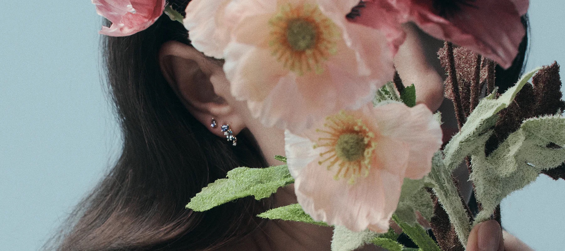 Woman with dark hair adorned with pink poppies and delicate earrings poses against a soft blue background in a beauty aesthetic.