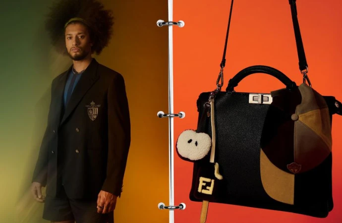 Adult male wearing blazer and shorts posing next to a black designer handbag with charm against split green and orange background.