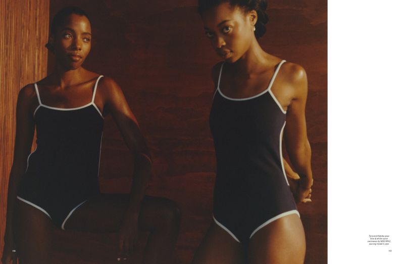 Two Black women model matching navy blue and white swimsuits in a warm, indoor setting with wood wall, photographed in medium shot.