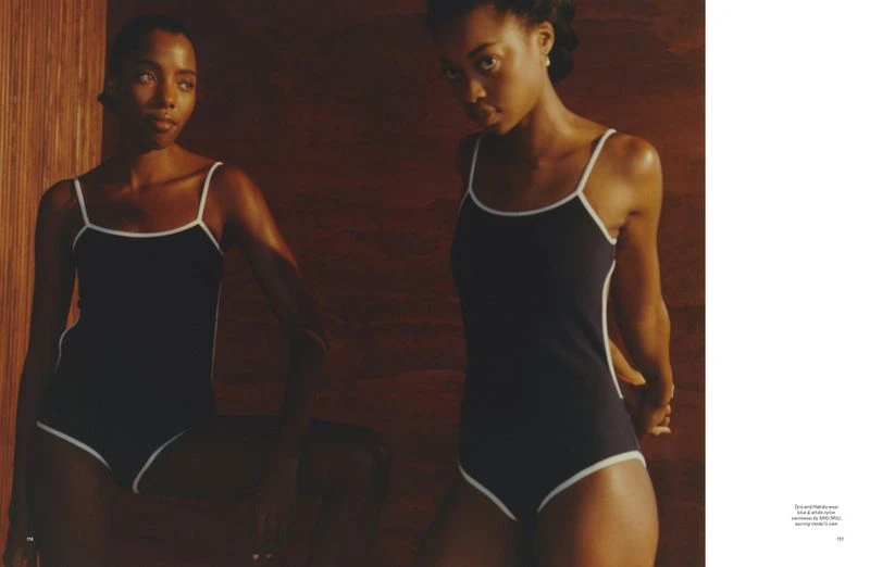 Two Black women model matching navy blue and white swimsuits in a warm, indoor setting with wood wall, photographed in medium shot.
