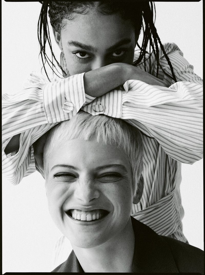 Two young adult females pose in a black-and-white studio portrait. The woman in the front has short blonde hair and a wide smile.