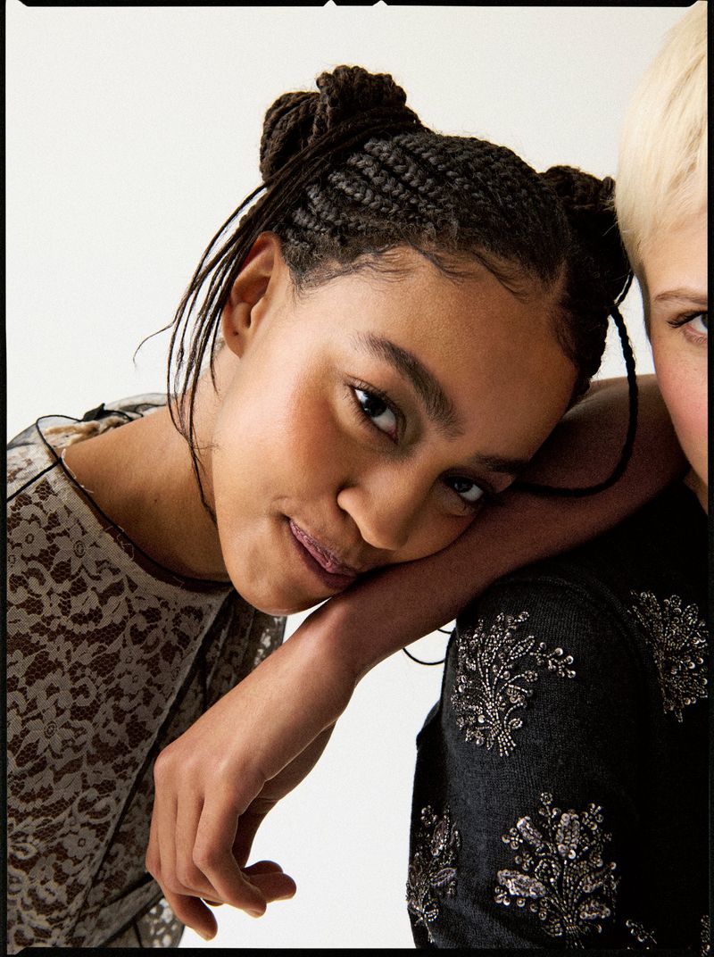 Close-up portrait of two young women with braided hair and fashionable tops leaning against each other in a bright indoor studio.