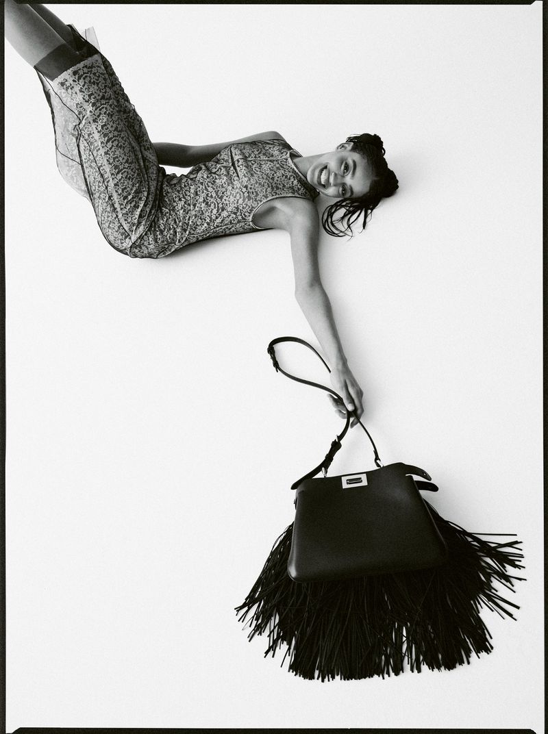 A woman in a patterned dress lies on a white surface, holding a black leather Fendi handbag with a fringe detail, creating a minimalist fashion image.