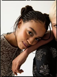 Close-up portrait of two female models, one with braided space buns resting head on another's shoulder against a white background.