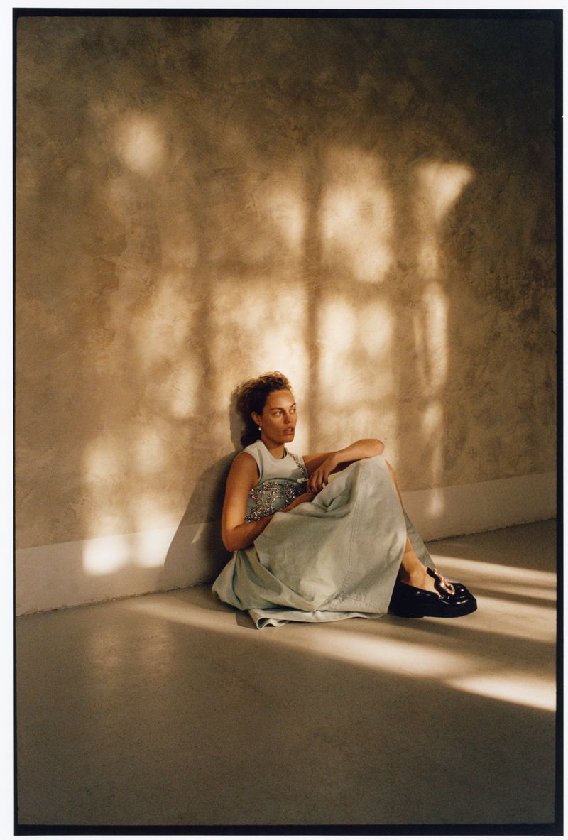 Woman in a sleeveless top and long skirt sitting indoors against a textured wall with shadows, holding a patterned clutch.