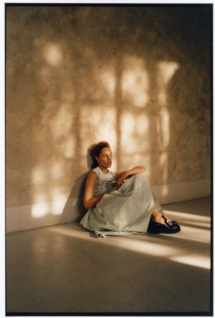 Woman in a sleeveless top and long skirt sitting indoors against a textured wall with shadows, holding a patterned clutch.