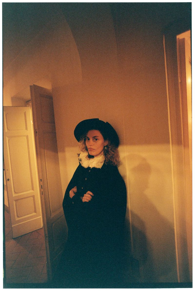 Stylish woman in a black cape and hat posing in an indoor setting with soft, warm lighting and neutral color palette. Fashion shot.
