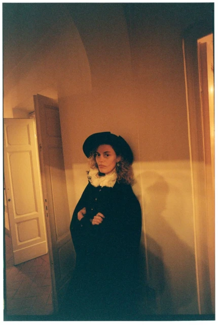 Stylish woman in a black cape and hat posing in an indoor setting with soft, warm lighting and neutral color palette. Fashion shot.