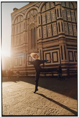 Female dancer strikes pose in plaza with a marble building and a crowd of people in the background, bathed in warm, late-day sunlight.
