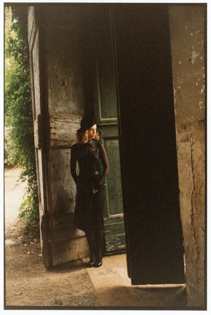 Adult female stands in front of a weathered green door, dressed in all black, wearing a hat in a garden setting, outdoor shot, medium framing.