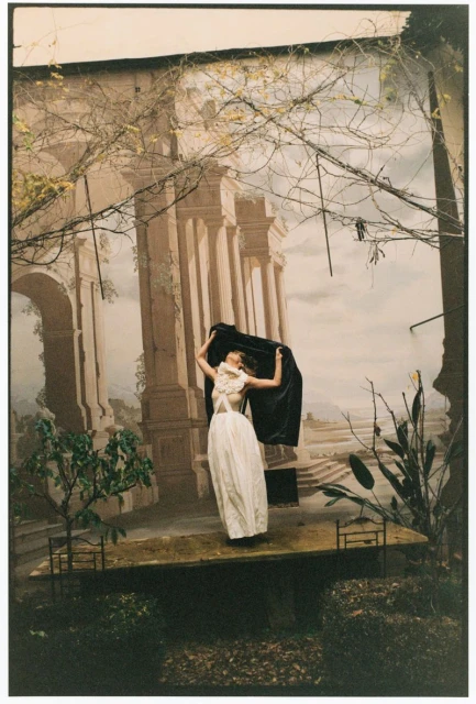 A woman in a white dress and black shawl poses against a backdrop with a painted colonnade, foliage, and sky in a fashion editorial shot.