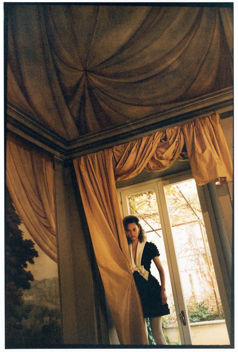 Woman in black dress with white trim and tights poses in an ornately decorated room with gold curtains and a painted ceiling.