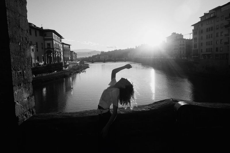 Adult female poses on a stone ledge overlooking a river with buildings in the background in a black and white photograph at sunset.