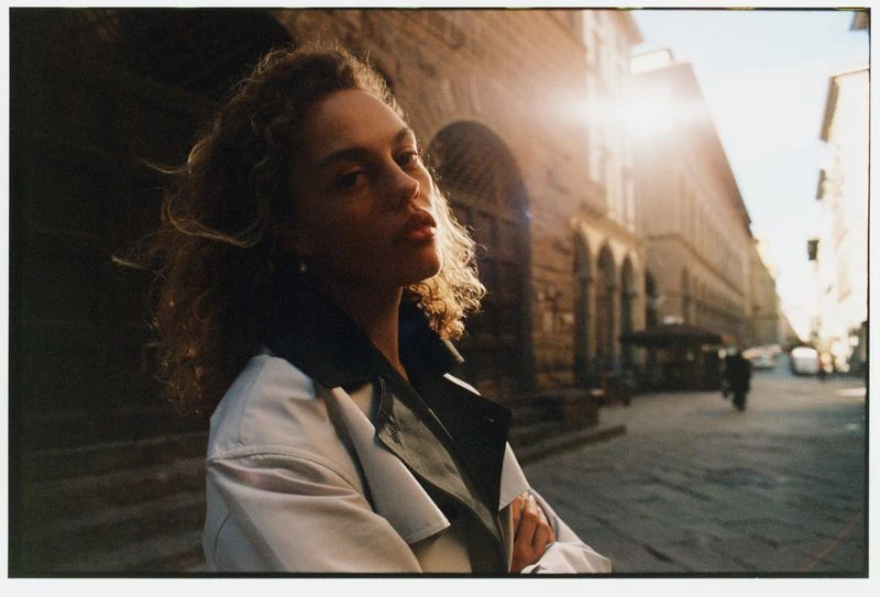Woman with curly hair poses in a trench coat on a cobblestone street, bathed in warm sunlight with blurred buildings in the background.