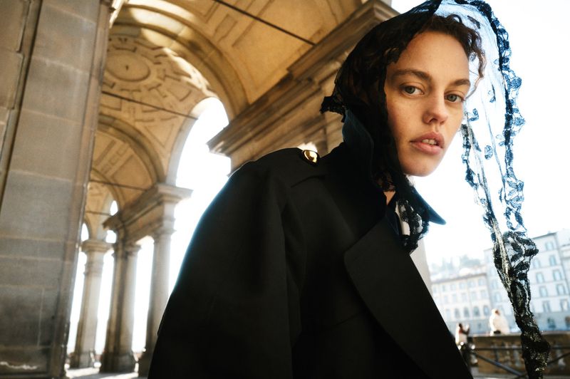 Woman in a black trench coat and lace head covering stands under stone arches in a low-angle fashion shot with blurred buildings.
