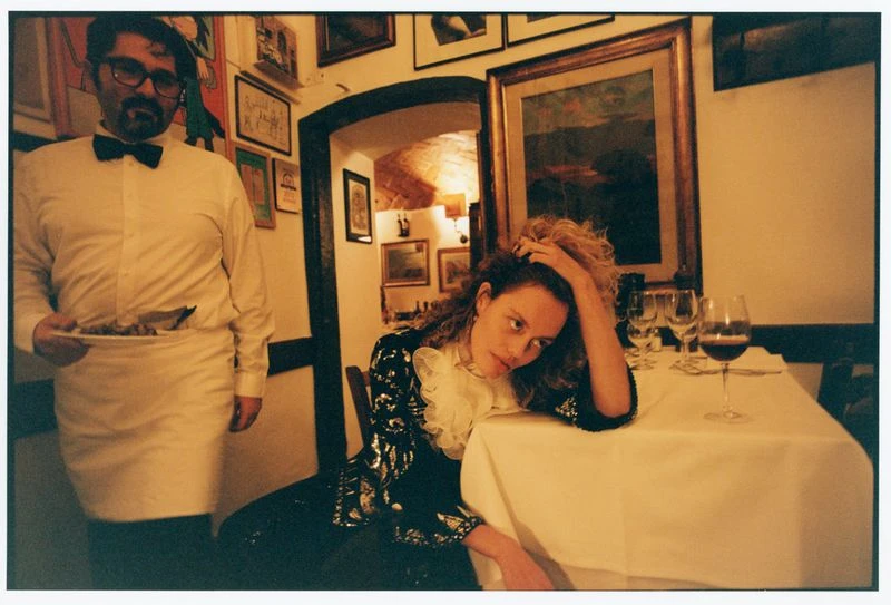 Woman at a table with a white tablecloth and wine glasses, served by a waiter in a white shirt and black bow tie, in a dimly lit restaurant.