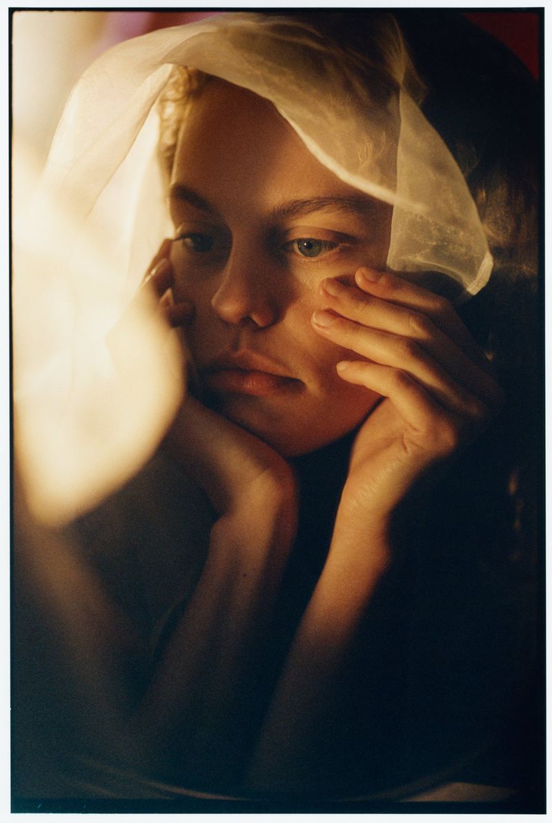 Close-up portrait of a woman with her hands framing her face and a translucent white fabric draped over her head in warm lighting.