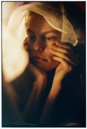 Close-up portrait of a woman with curly hair wearing a sheer white fabric draped over her head, resting her chin on her hands
