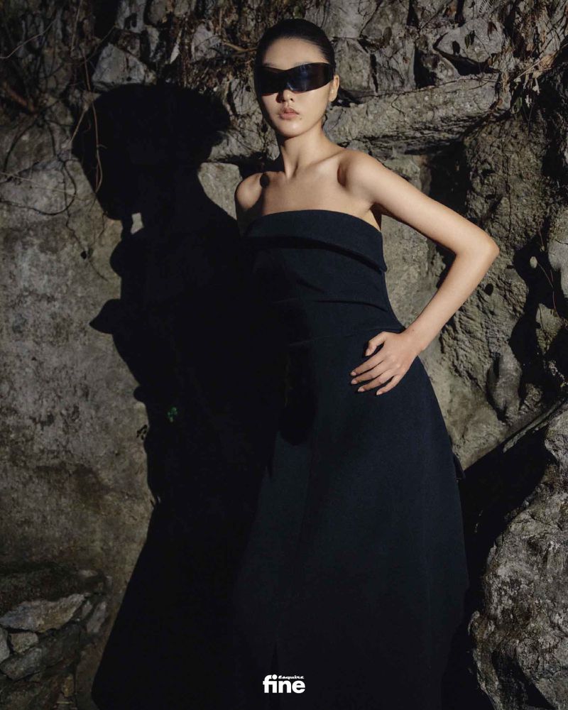 A fashionable woman poses in a black strapless dress and futuristic glasses against a textured stone wall in moody, shadowed lighting.