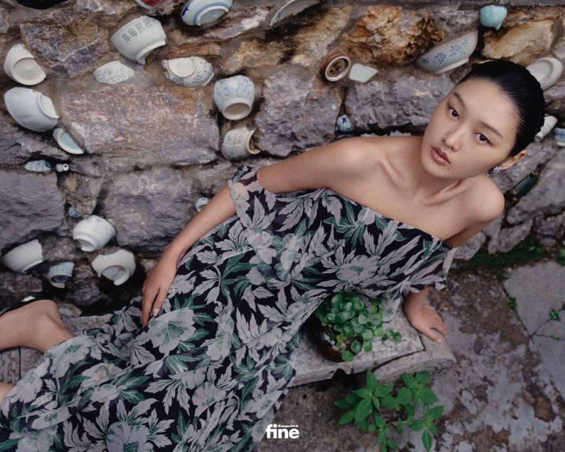 Adult female model in a floral dress posing near a rustic stone wall with embedded porcelain bowls, creating a blend of fashion and culture.