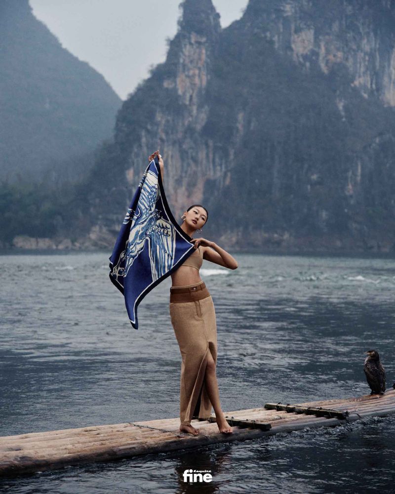 Asian woman poses on a wooden platform in a river holding a blue and white scarf, with mountains visible in the background.