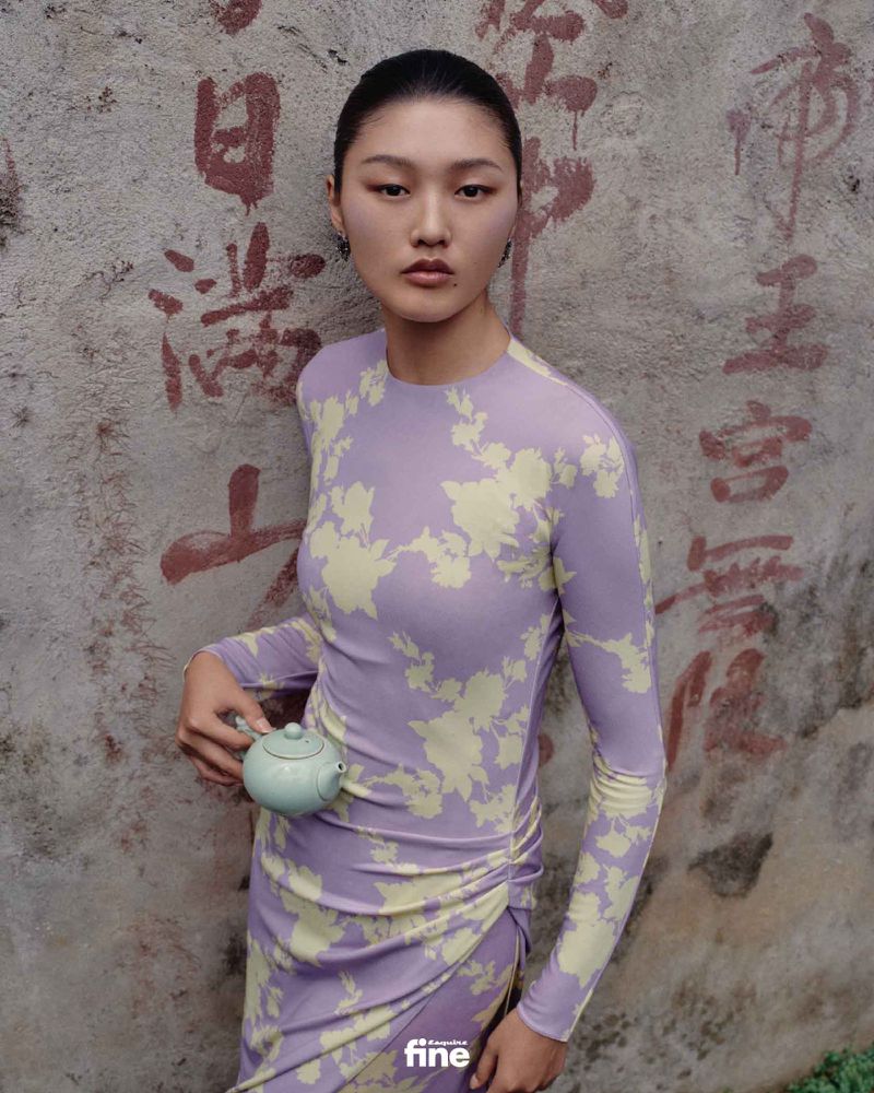 Asian woman in a patterned dress poses with a light green teapot against a concrete wall with faded, dark red Chinese script.