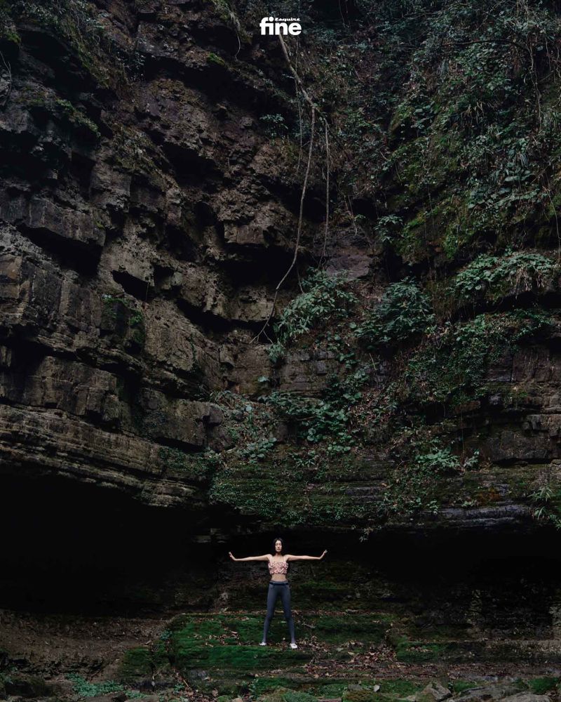 Adult female stands with arms outstretched in front of a rock cliff covered with moss and vegetation, wearing athletic wear, outdoors.